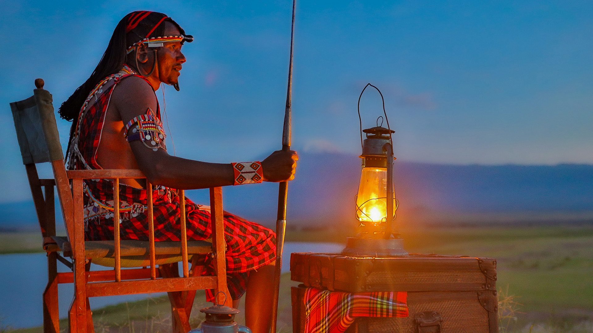 Maasai guide seated at a sundowner setup with lantern and trunk at dusk on the savanna