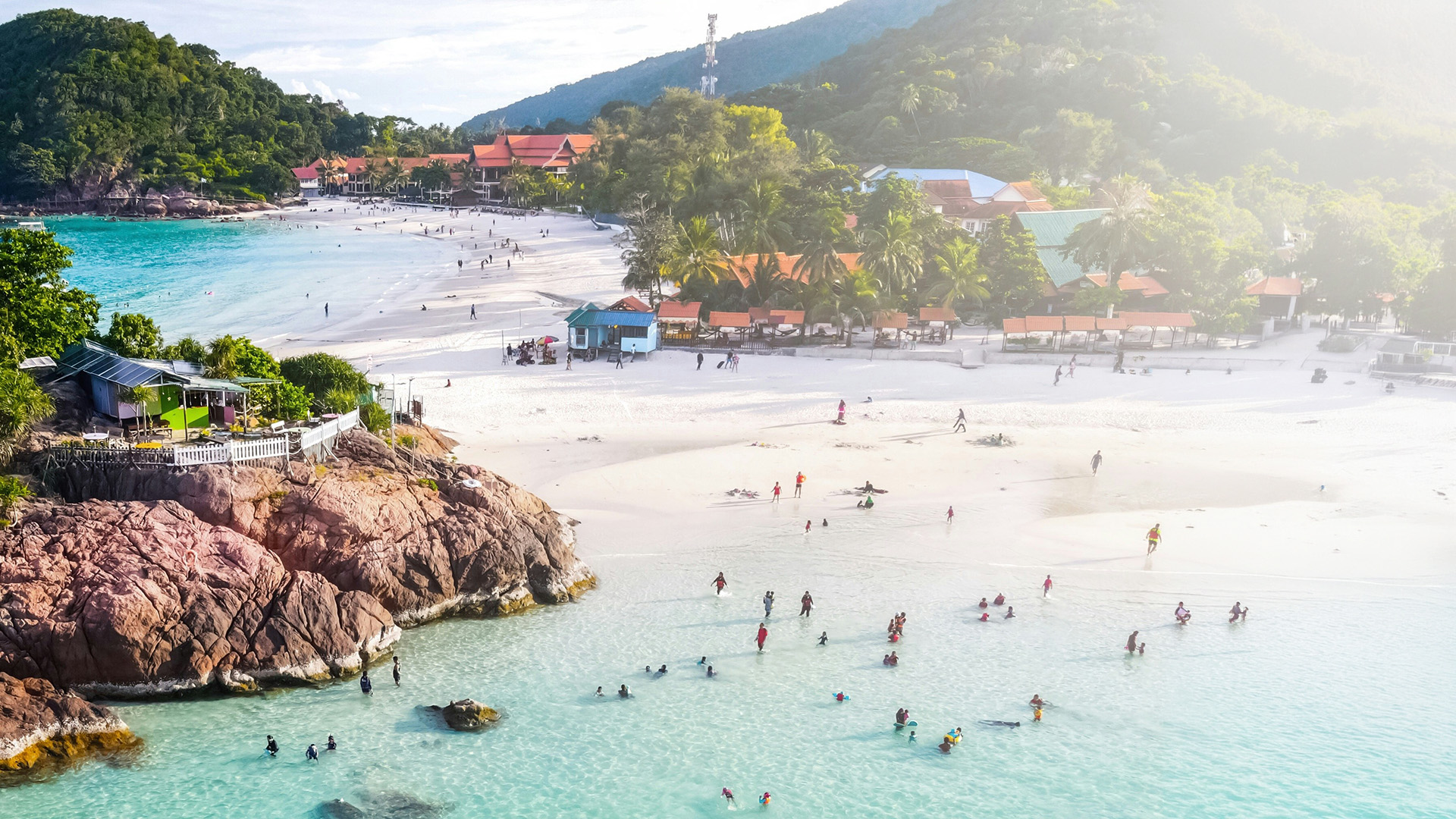 Aerial view of a busy tropical beach resort with turquoise water and forested hills in Malaysia
