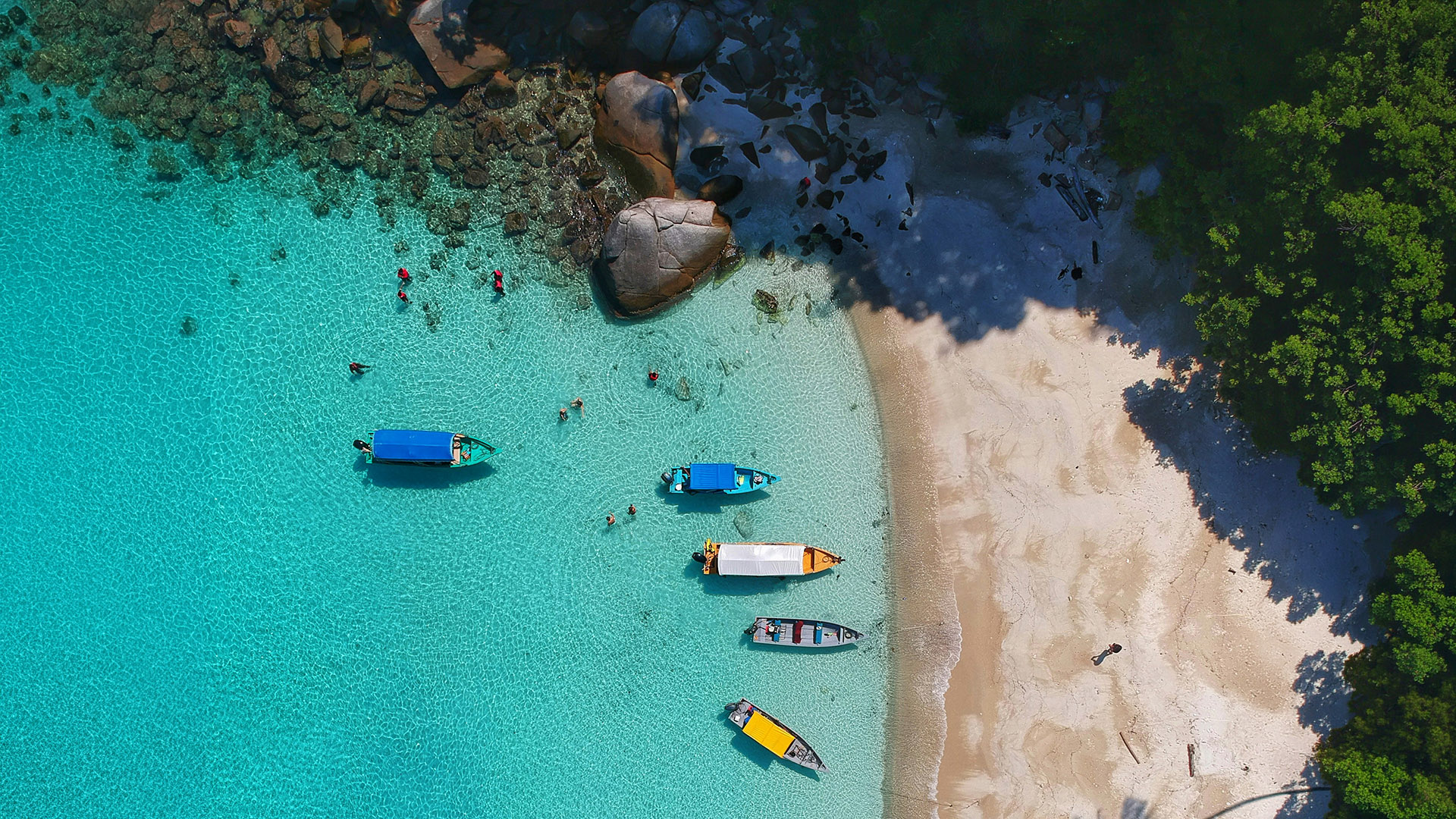 Overhead view of boats in clear turquoise water beside a white-sand beach and tropical forest